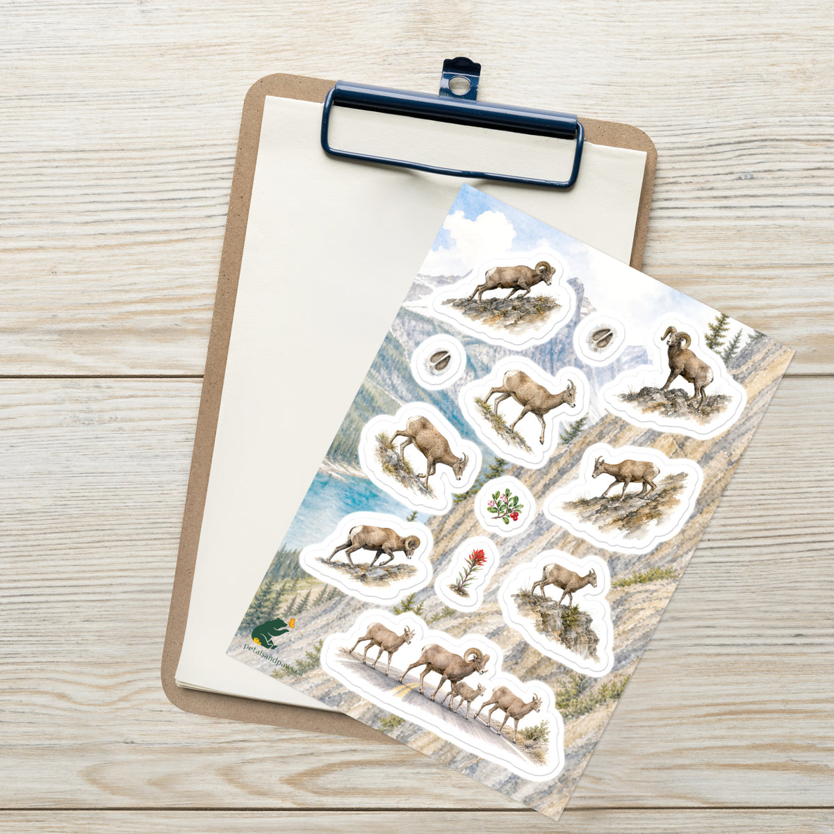 A sheet of kiss-cut stickers featuring illustrated bighorn sheep on the move. set against Rocky Mountain scenery, placed on top of an open clipboard with a blank sheet on a light wood tabletop.
