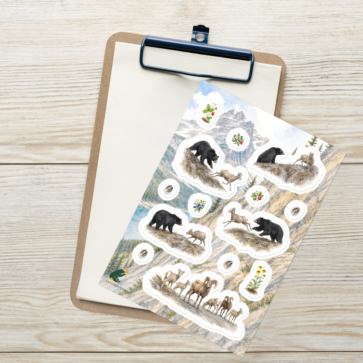 A sheet of kiss-cut stickers featuring illustrated black bear and bighorn sheep, roadway herd and botanicals, set against Rocky Mountain scenery, placed on top of an open clipboard with a blank sheet on a light wood tabletop.