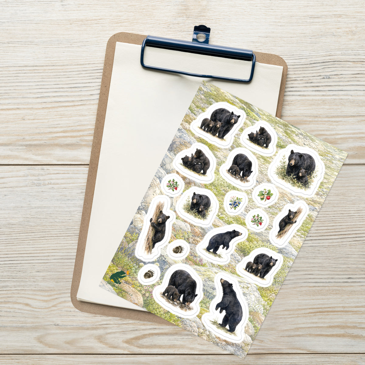 A sheet of kiss-cut stickers featuring illustrated black bear families and mountain terrain, placed on top of an open clipboard with a blank sheet on a light wood tabletop.