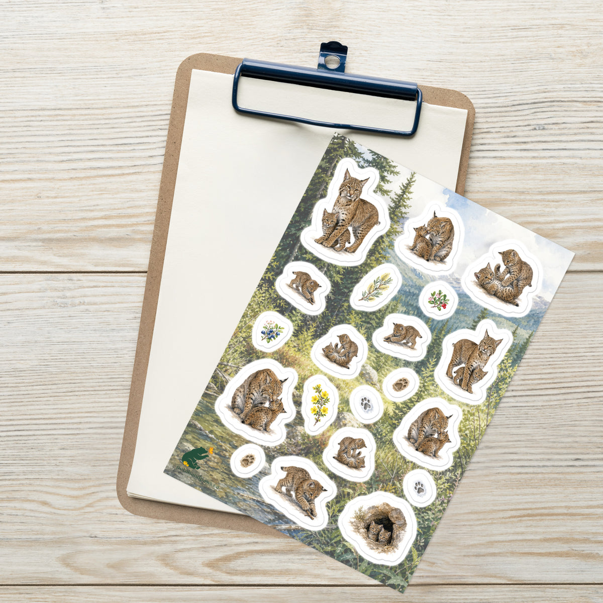 A sheet of kiss-cut stickers featuring illustrated bobcat mothers and kittens, set against Rocky Mountain foothills scenery, and placed on top of an open clipboard with a blank sheet on a light wood tabletop.