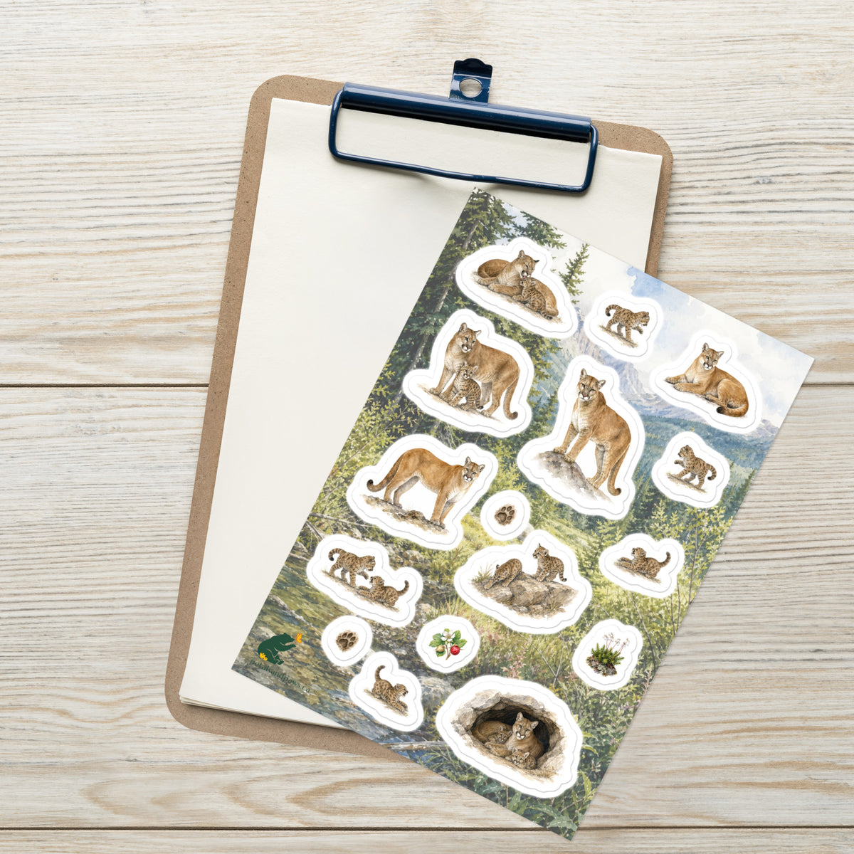 A sheet of kiss-cut stickers featuring illustrated mountain lion mothers and cubs, set against Rocky Mountain foothills scenery, and placed on top of an open clipboard with a blank sheet on a light wood tabletop.