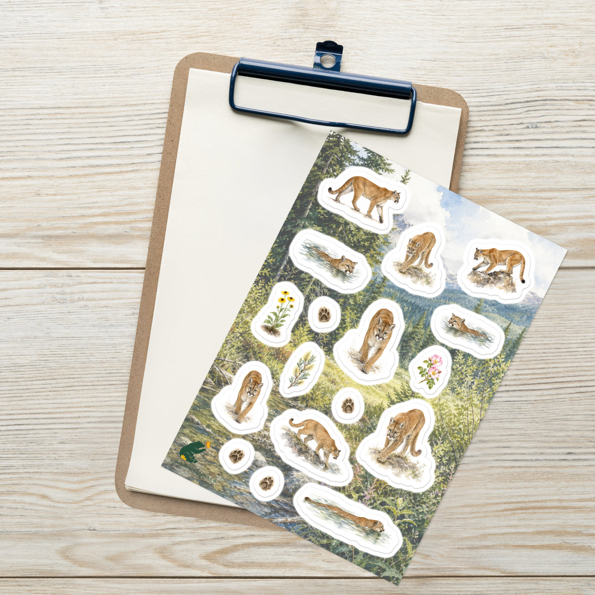 A sheet of kiss-cut stickers featuring illustrated traveling cougars and botanicals, set against Rocky Mountain foothills scenery, placed on top of an open clipboard with a blank sheet on a light wood tabletop.