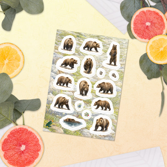A sheet of kiss-cut stickers featuring illustrated grizzly bears on the move, with a background of rocky mountain icefields terrain, placed next to citrus slices on a warm-colored background.
