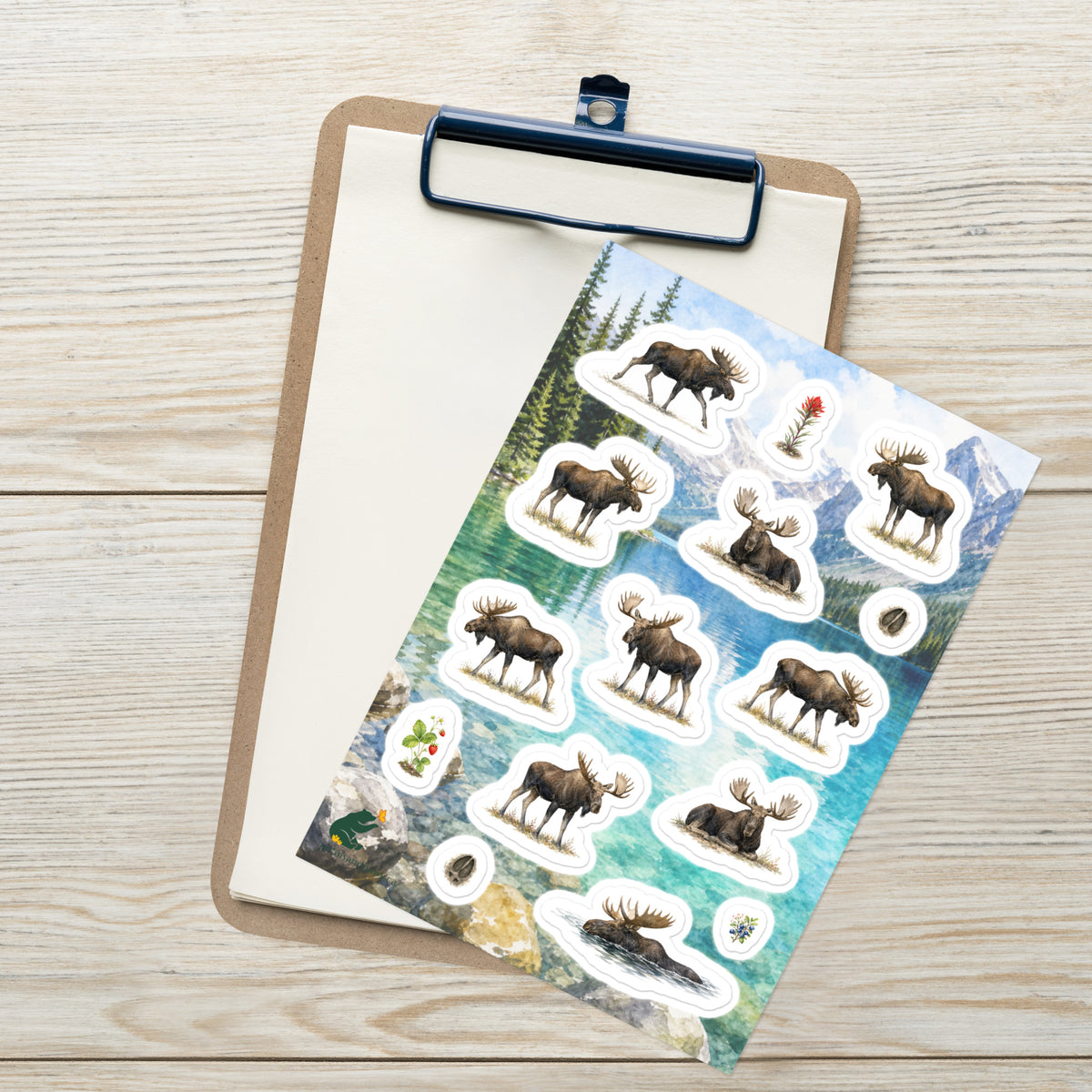 A sheet of kiss-cut stickers featuring illustrated mature bull moose, set against Rocky Mountain lake scenery, and placed on top of an open clipboard with a blank sheet on a light wood tabletop.