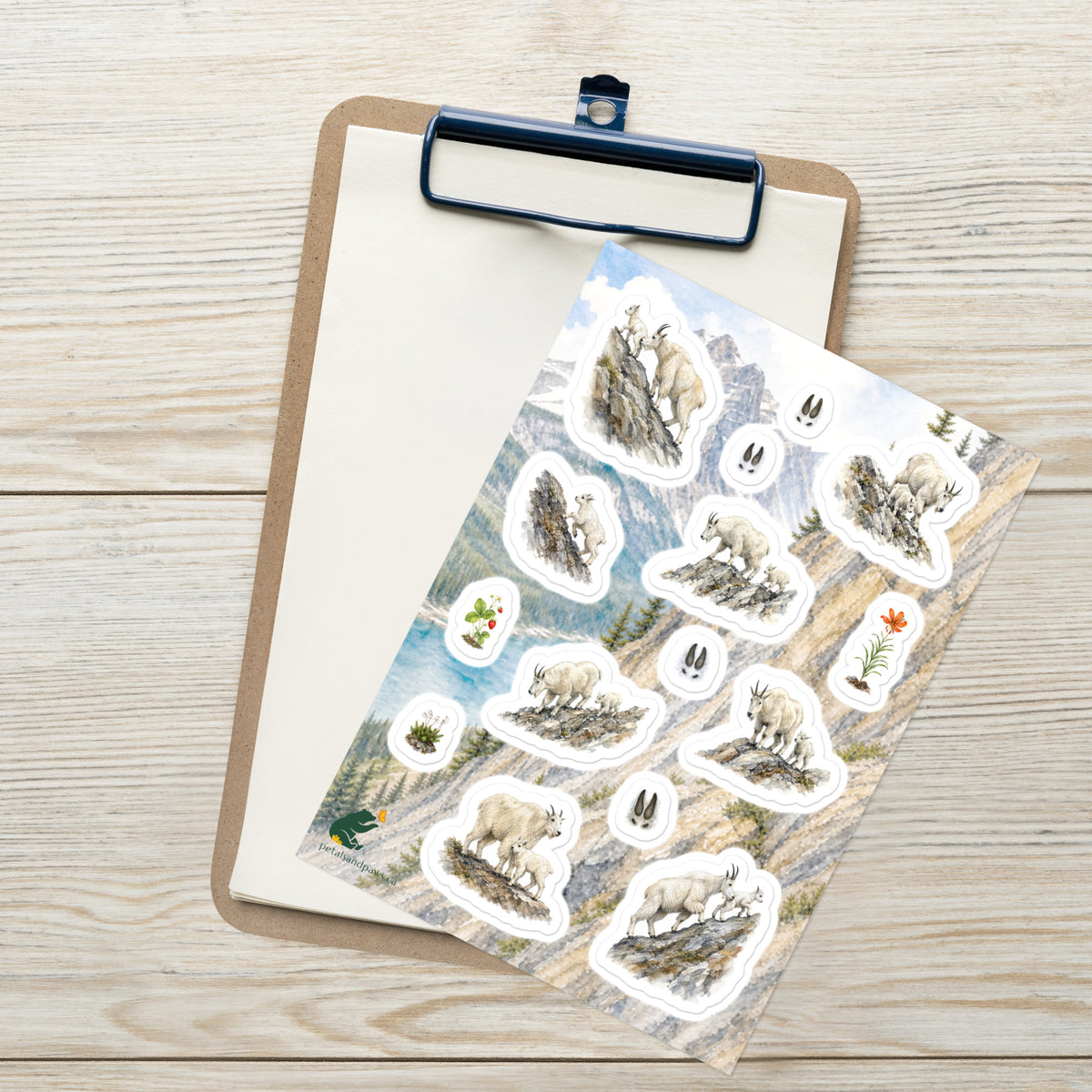 A sheet of kiss-cut stickers featuring illustrated mountain goat nannies and kids, set against Rocky Mountain scenery, placed on top of an open clipboard with a blank sheet on a light wood tabletop.