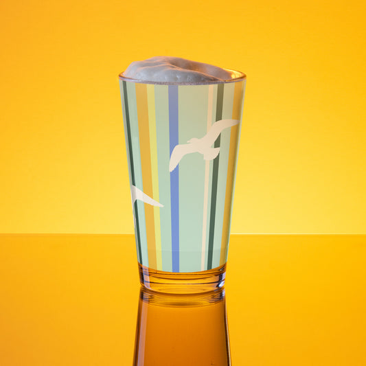 In-action mockup of Petals & Paws Cove Pint Glass, featuring seagull design with teal striped pattern. The glass, surrounded by orange glow, is holding beer with frothy foam.