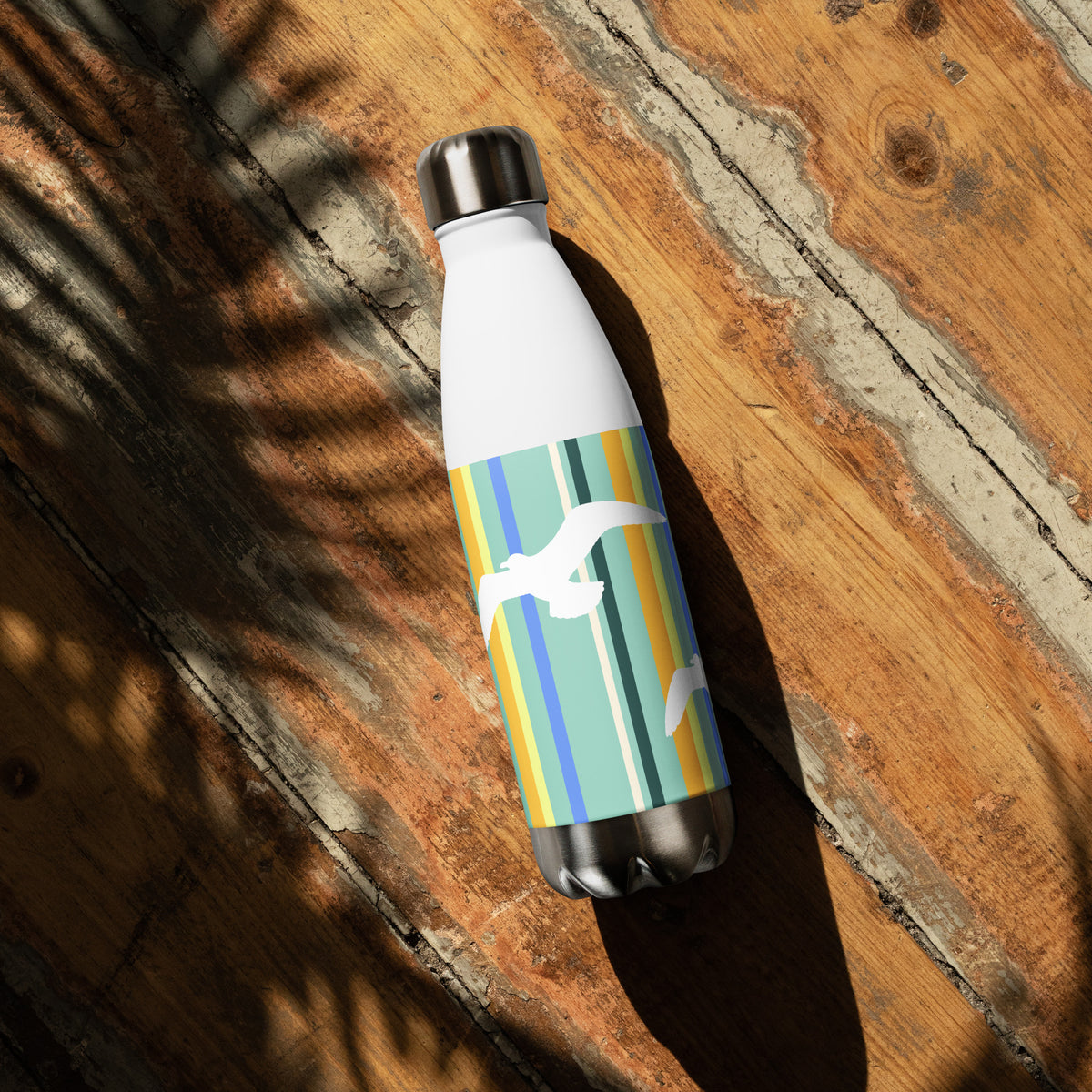 A 17 oz Petals & Paws Cove Water Bottle laying down on a sunny wooden deck with the shadow of a plant. This bottle features seagull design with teal striped pattern.
