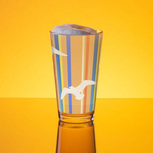 In-action mockup of Petals & Paws Drift Pint Glass, featuring seagull design with orange striped pattern. The glass, surrounded by orange glow, is holding beer with frothy foam.