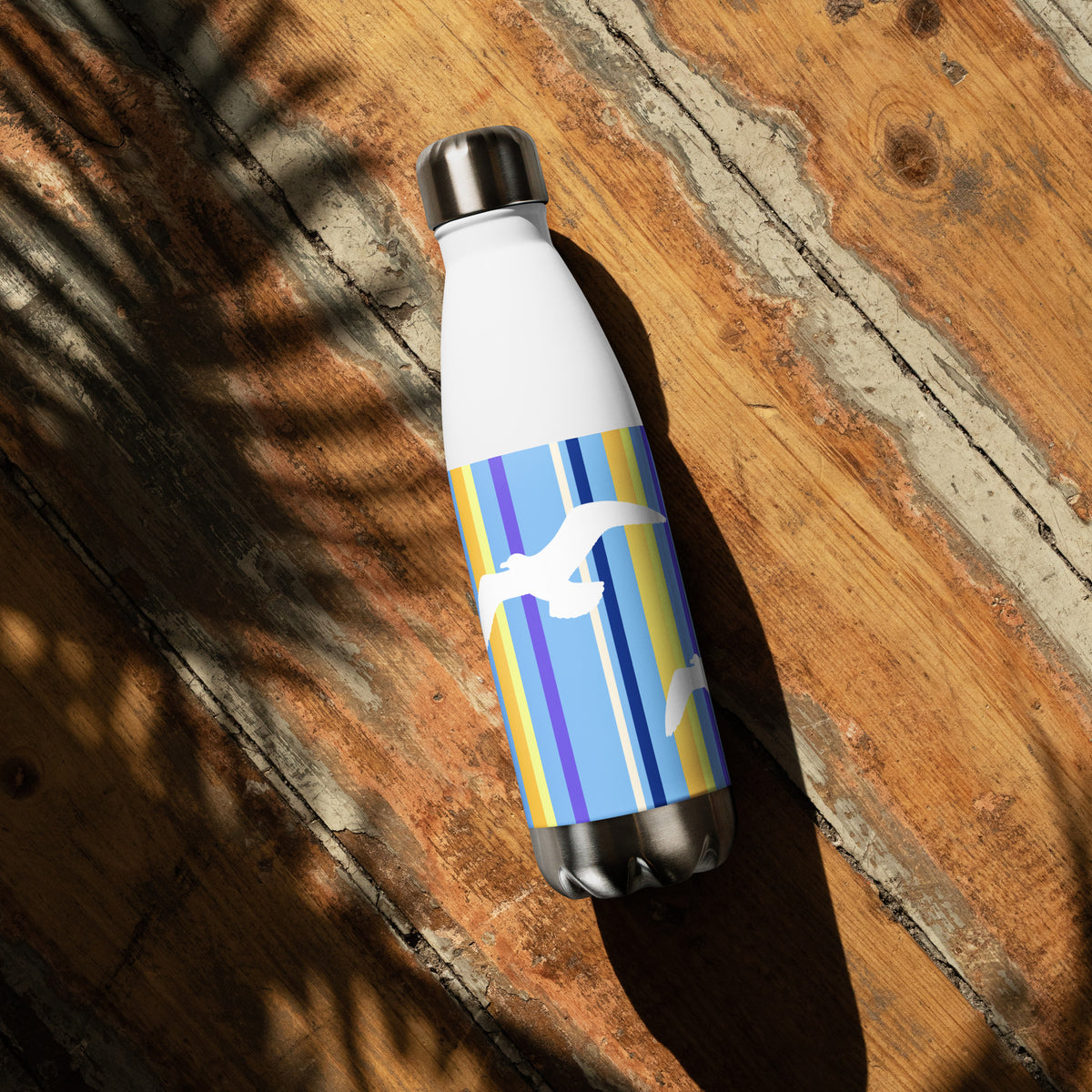A 17 oz Petals & Paws Shoreline Water Bottle laying down on a sunny wooden deck with the shadow of a plant. This bottle features seagull design with blue striped pattern.
