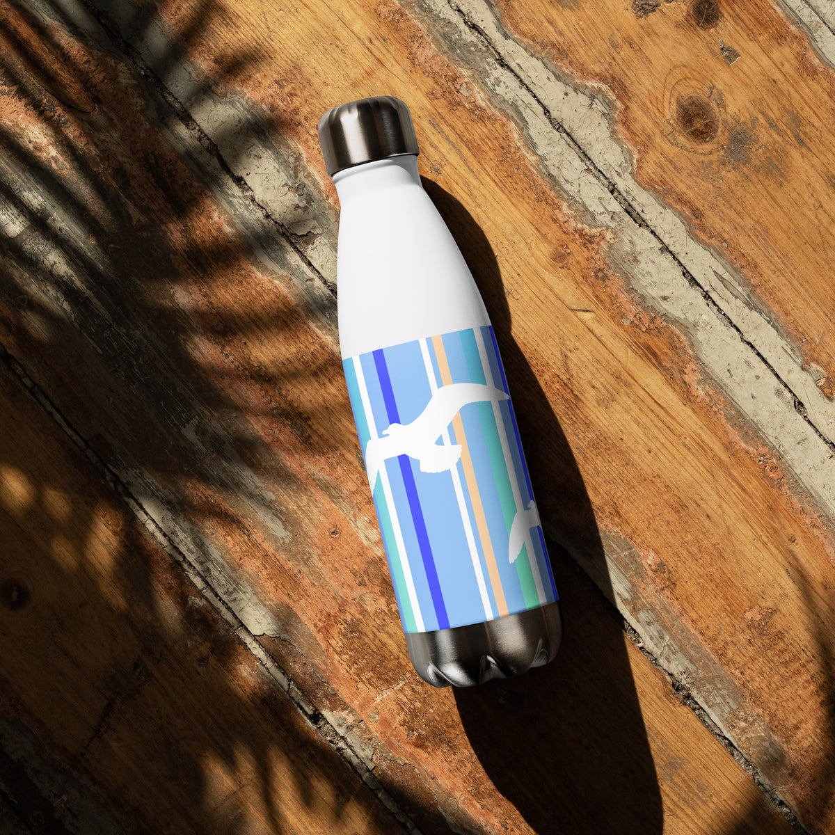 A 17 oz Petals & Paws Tide Water Bottle laying down on a sunny wooden deck with the shadow of a plant. This bottle features seagull design with blue striped pattern.