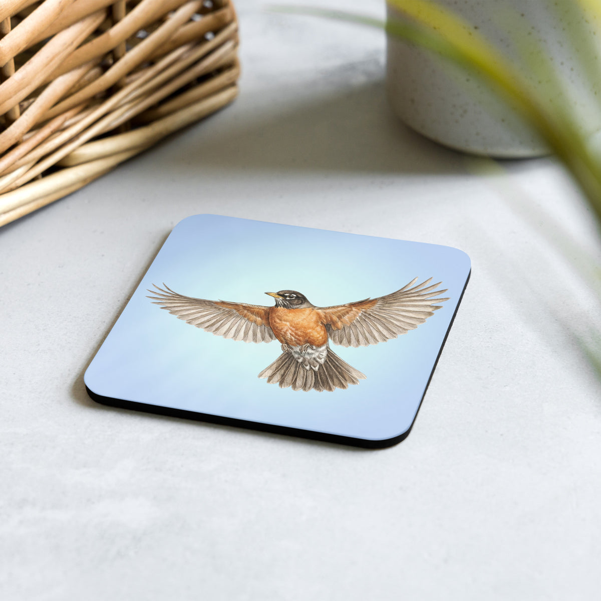 Product mockup of American Robin Coaster set on a desk with a woven basket and clay mug. Robin is flying against a clear blue sky.