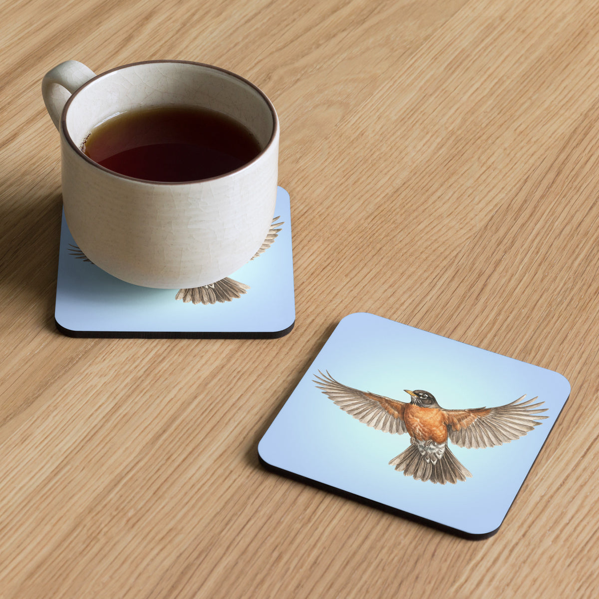 Product mockup of two American Robin Coasters, one with a mug of tea sitting on it, and the other set beside, both on a faux-wood surface. Robin is flying against a clear blue sky.