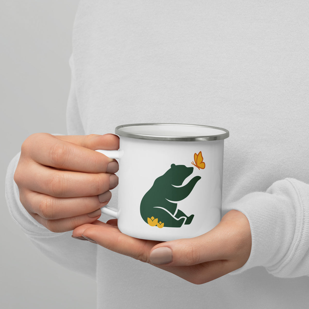 A woman holds the handle of 12 oz Beary Good Enamel Mug for left-hand orientation in her right hand.  This reveals Petals & Paws green bear graphic with yellow butterfly and flowers.