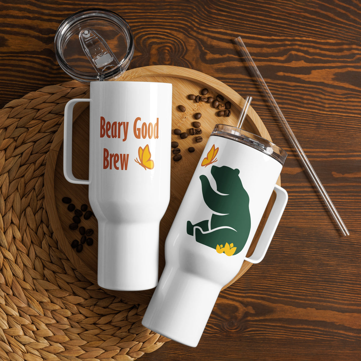 Two 40 oz Beary Good Travel right-oriented mugs laying on a wooden platter with some scattered coffee bean. One shows the Petals & Paws green graphic bear that faces the drinker, while the other show the "Beary Good Brew" text that faces out and the straw that comes with this 40 oz version.