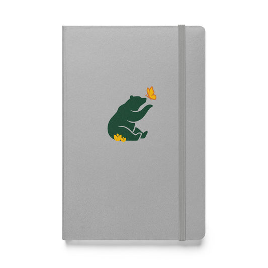 Flat-lay of gray (grey) notebook with green Petals & Paws bear graphic, sitting in yellow flowers with paw extended to yellow and orange butterfly.