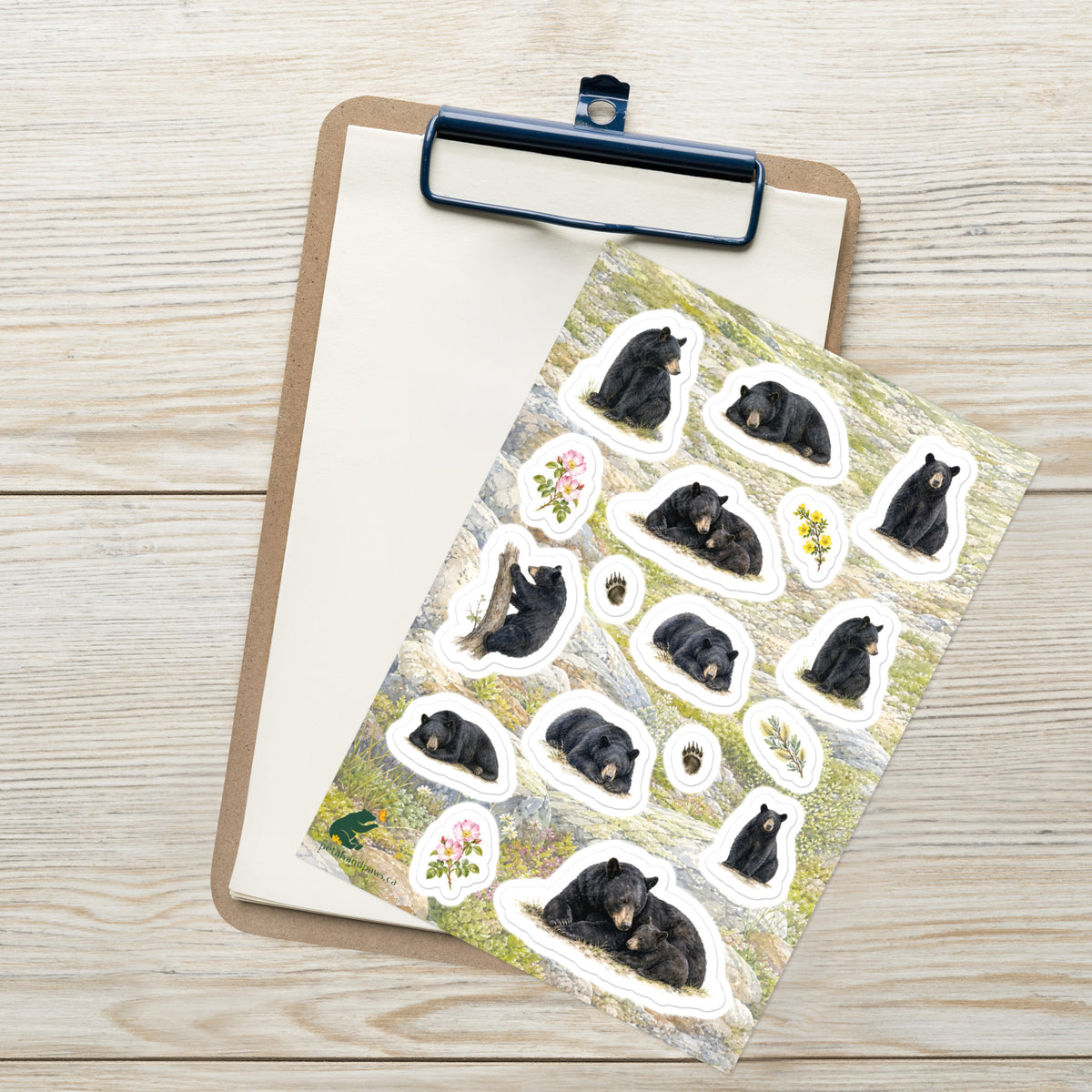 A sheet of kiss-cut stickers featuring illustrated black bears resting and mountain terrain, placed on top of an open clipboard with a blank sheet on a light wood tabletop.