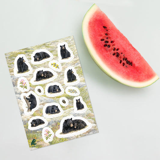 A sheet of kiss-cut stickers featuring illustrated black bears resting, with a background of rocky mountain icefields terrain, placed next to a slice of watermelon.