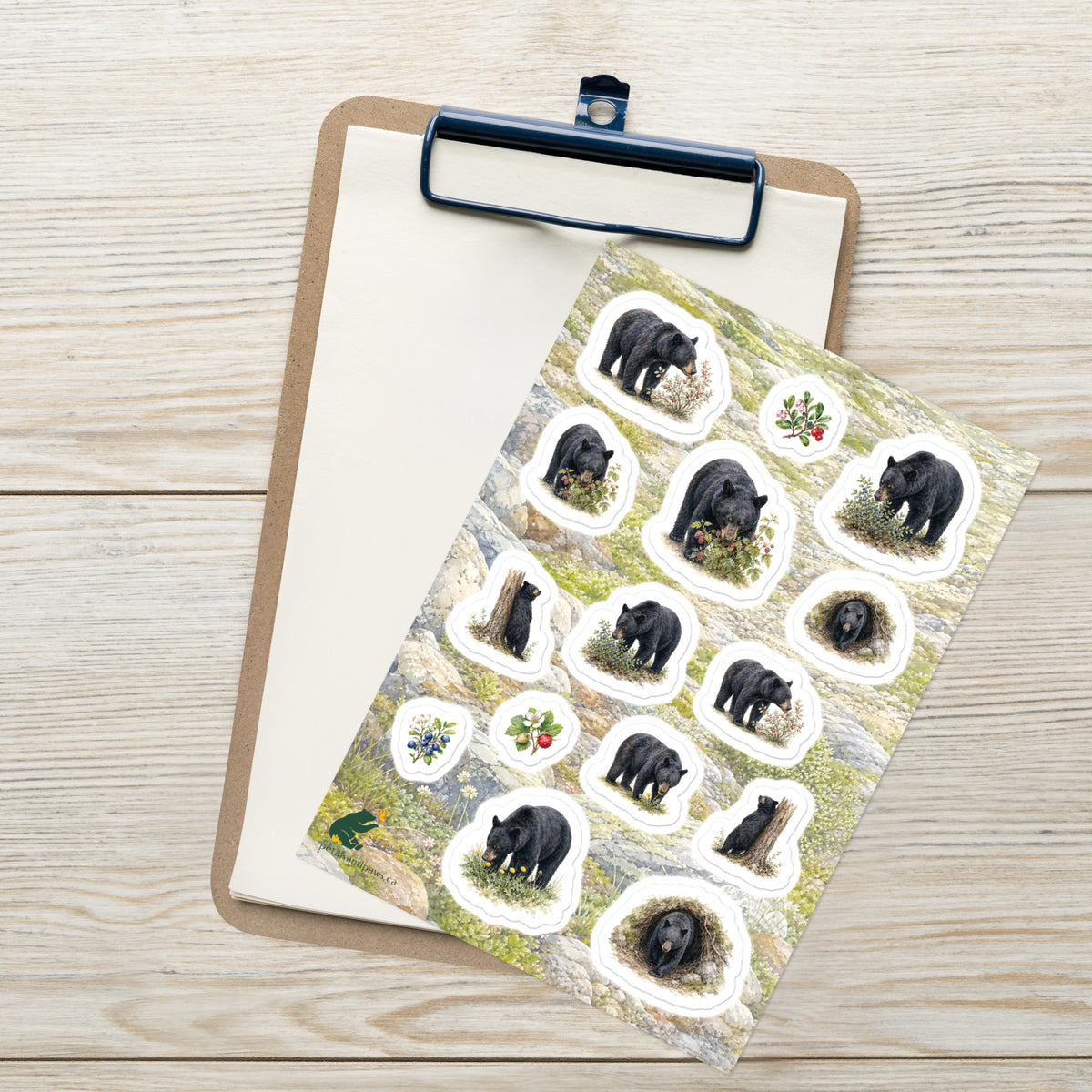 A sheet of kiss-cut stickers featuring illustrated black bears foraging and mountain terrain, placed on top of an open clipboard with a blank sheet on a light wood tabletop.