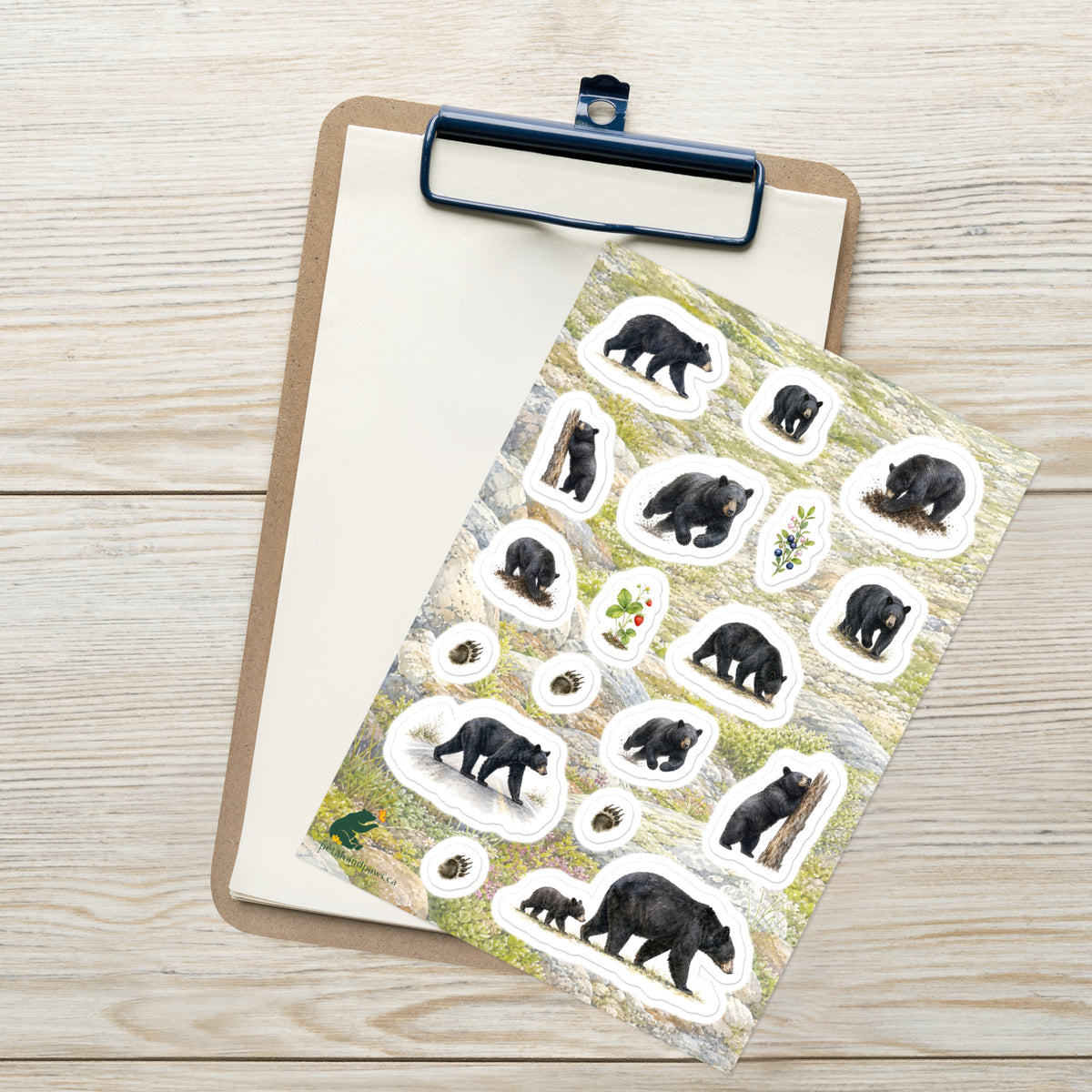 A sheet of kiss-cut stickers featuring illustrated black bears on the move and mountain terrain, placed on top of an open clipboard with a blank sheet on a light wood tabletop.