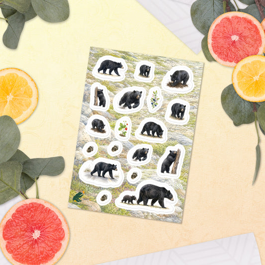 A sheet of kiss-cut stickers featuring illustrated black bears on the move, with a background of rocky mountain icefields terrain, placed next to citrus slices on a warm-colored background.
