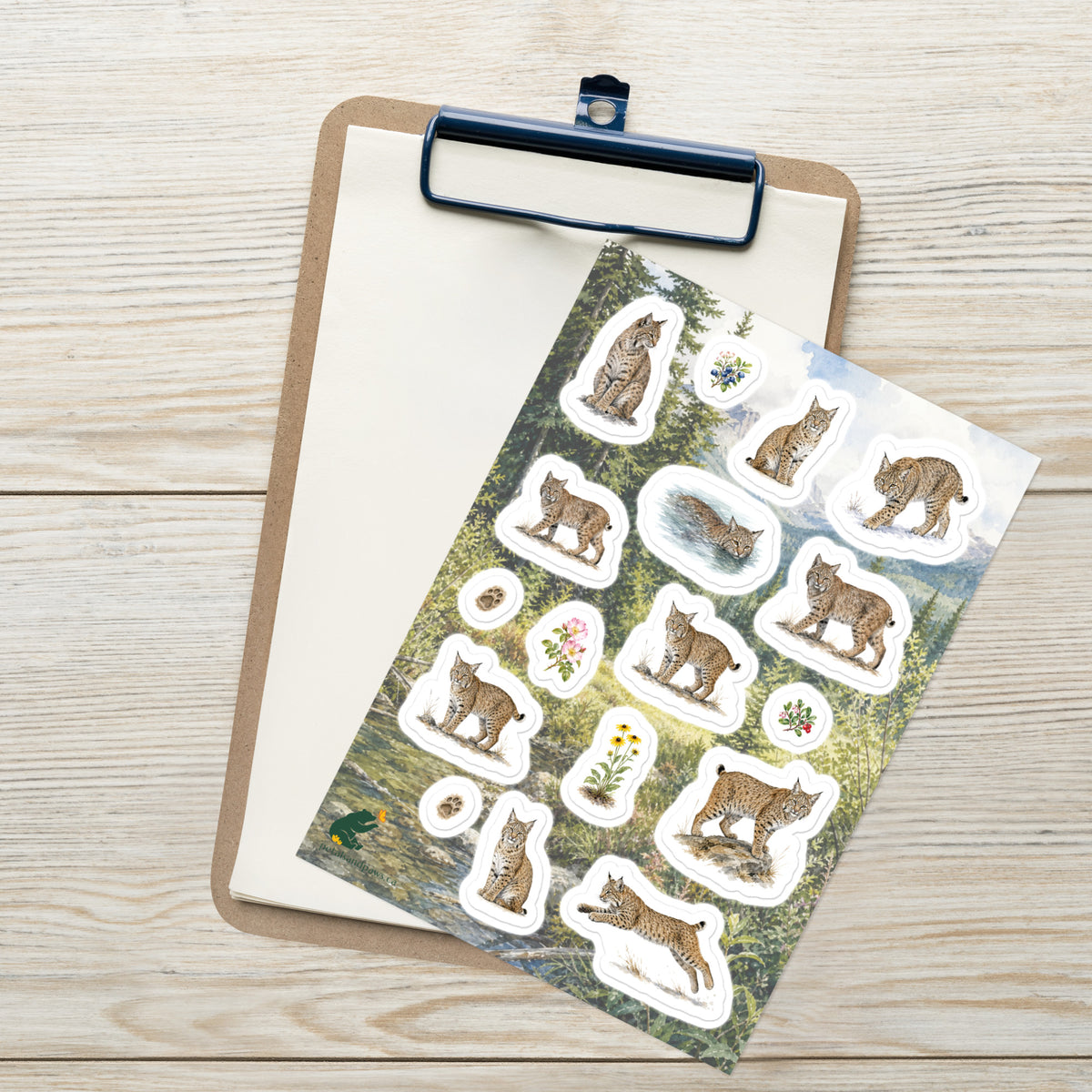 A sheet of kiss-cut stickers featuring illustrated bobcats, set against Rocky Mountain foothills scenery, placed on top of an open clipboard with a blank sheet on a light wood tabletop.