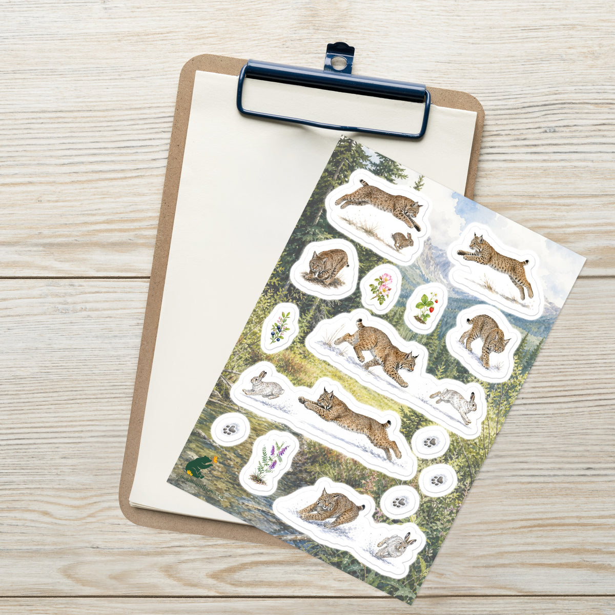 A sheet of kiss-cut stickers featuring illustrated bobcats hunting, set against Rocky Mountain foothills scenery, placed on top of an open clipboard with a blank sheet on a light wood tabletop.