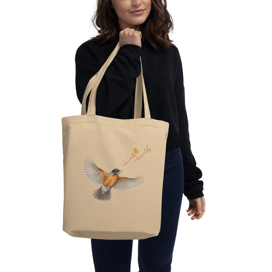A woman holding the Cheerily Robin Eco Tote Bag, with a flying robin, singing, "cheerio cheerily" set against the ecru 100% organic cotton.  Print is on one side. The Petals & Paws orange and yellow butterfly accents the songster's text.