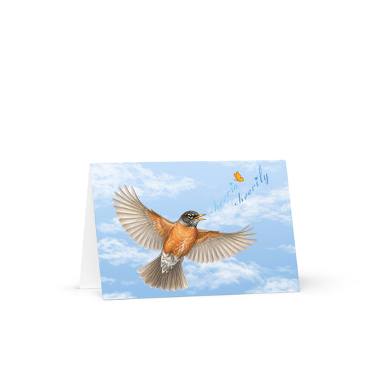 Product mockup of the Cheerily Robin Greeting Card, with a flying robin, singing, "cheerio cheerily" against a fair blue sky and fluffy cloud background. The Petals & Paws orange and yellow butterfly accents the songster's text.