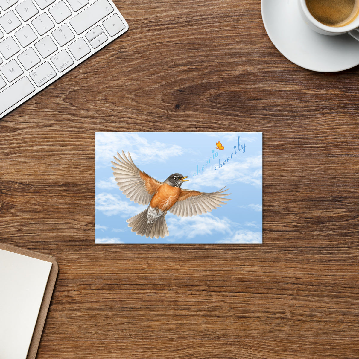 Product placement of the Cheerily Robin Greeting Card, with a flying robin, singing, "cheerio cheerily" against a fair blue sky and fluffy cloud background on an office desking. A keyboard, coffee cup and clipboard are partially visible.