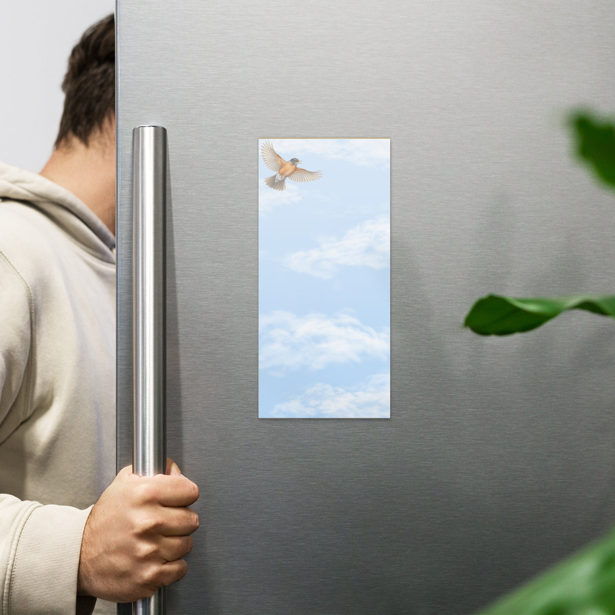 Cheerily Robin magnetic notepad, 4.25″ × 9″ (10.8 cm × 22.9 cm) displayed on fridge.  Overall design of fair blue sky with fluffy white clouds and a robin flying in the upper left corner.