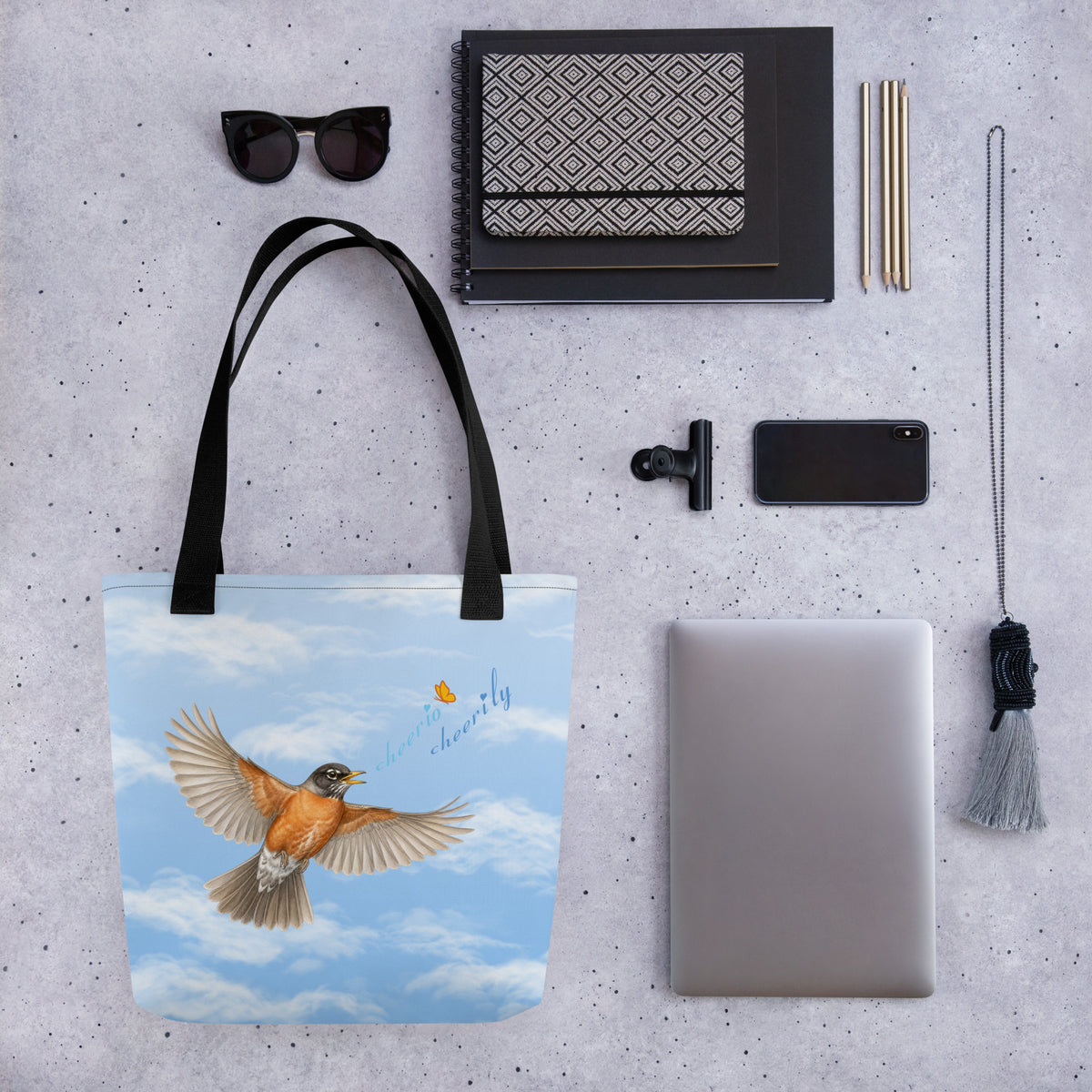 Product mockup of Cheerily Robin Tote Bag with various items that can fit inside set out beside it:  small laptop, decorative chain, cell phone, pencils, bulldog clip, a stack of notebooks, and sunglasses.