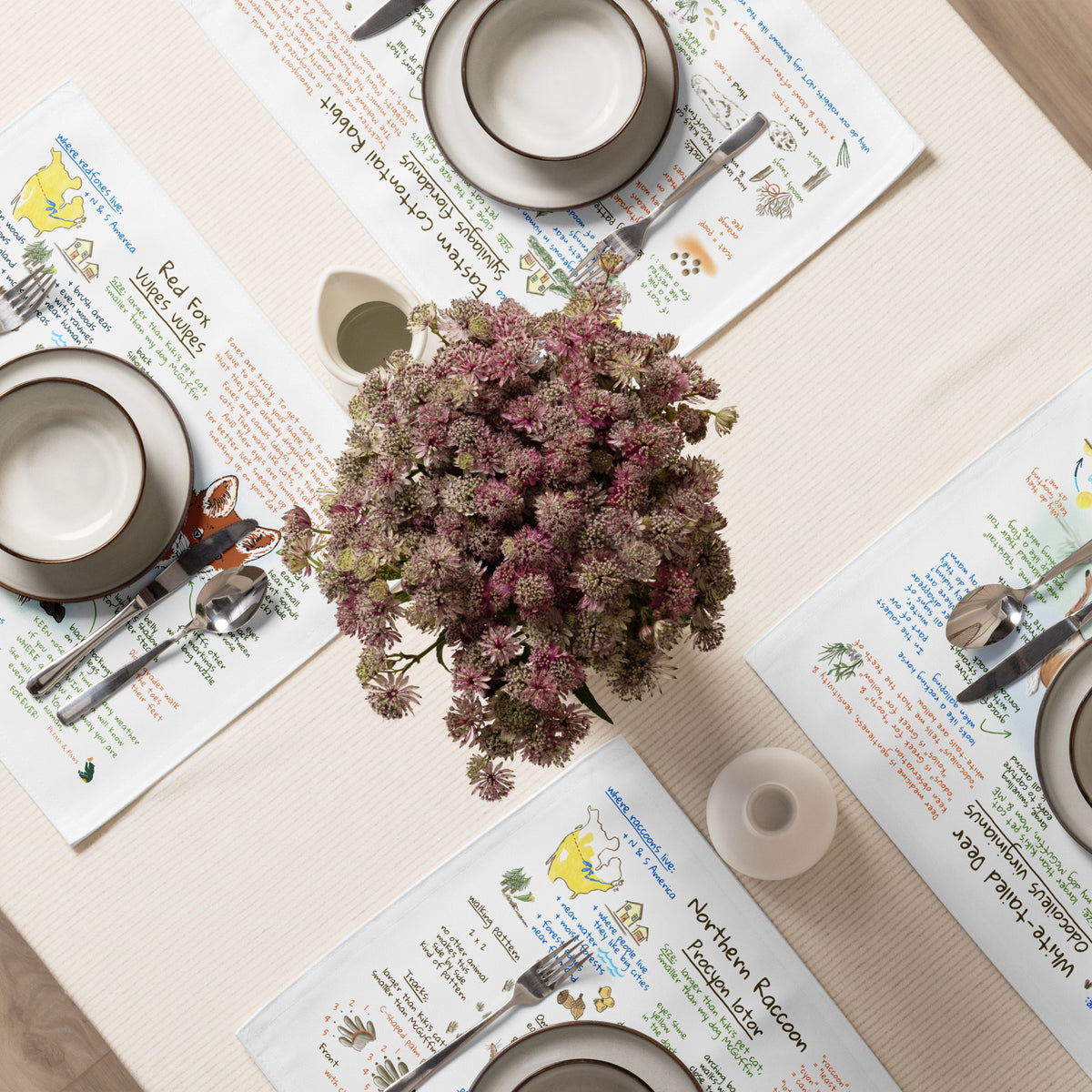 Overhead view of a table set with plates, bowls and utensils on top of Ezra's Bestiary Field Guide placemats. Cut flowers stand in the middle.