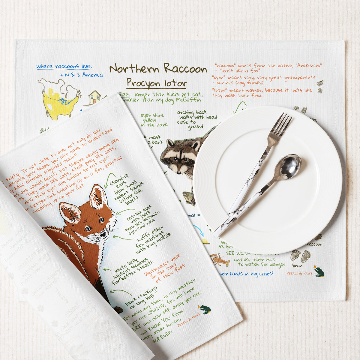 Ezra's Red Fox and Northern Raccoon  Bestiary Field Guide Placemats are featured on a cream-white background with a plate, fork and spoon.