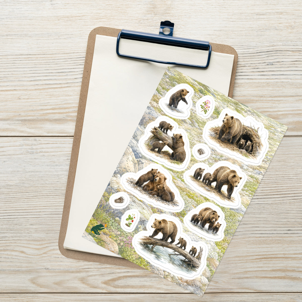 A sheet of kiss-cut stickers featuring illustrated grizzly bear families and mountain terrain, placed on top of an open clipboard with a blank sheet on a light wood tabletop.