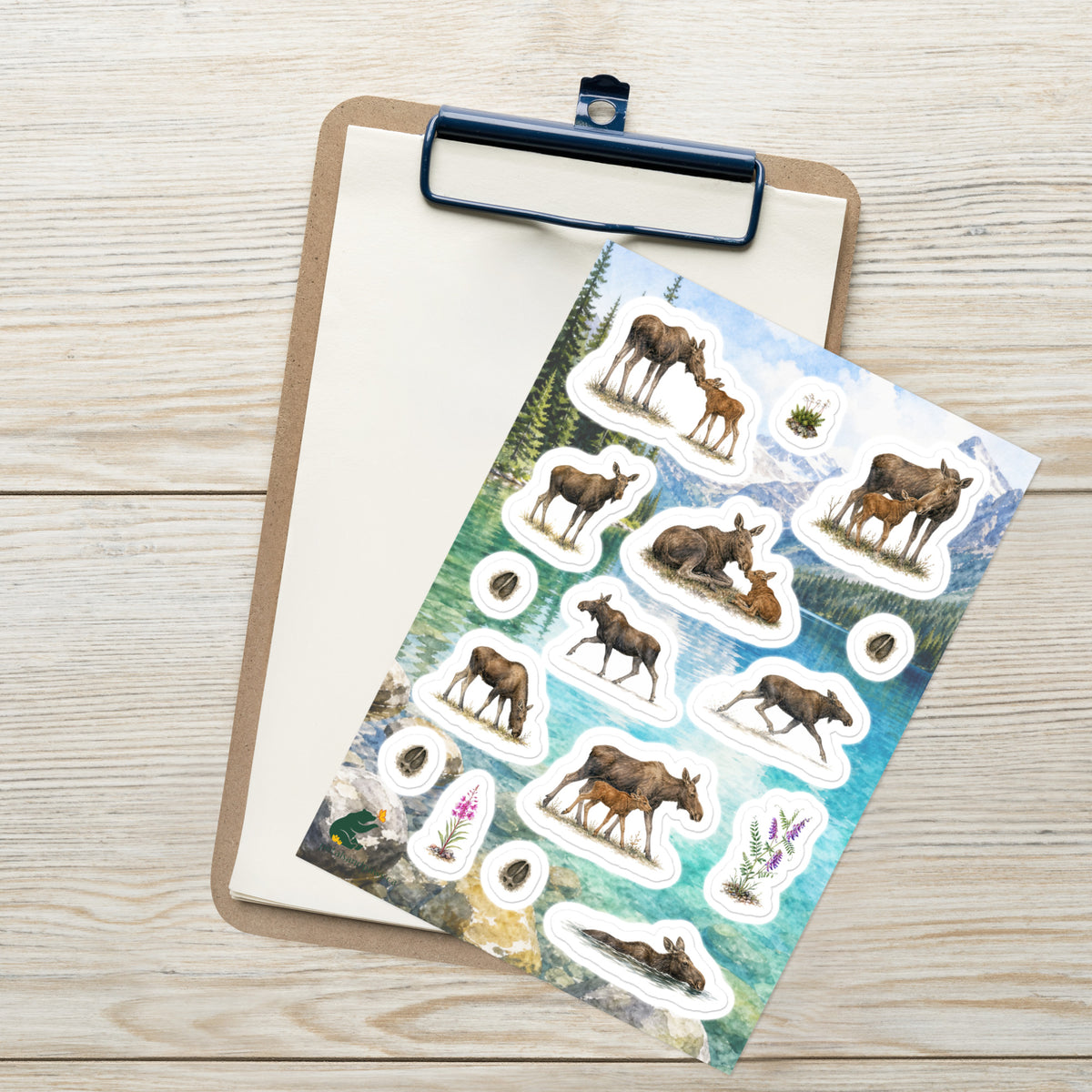 A sheet of kiss-cut stickers featuring illustrated moose cows and calves, set against Rocky Mountain lake scenery, and placed on top of an open clipboard with a blank sheet on a light wood tabletop.