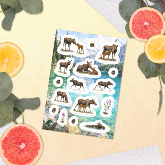 A sheet of kiss-cut stickers featuring illustrated moose cows and calves, set against a Rocky Mountain lake scene, and placed next to a slice of watermelon. 