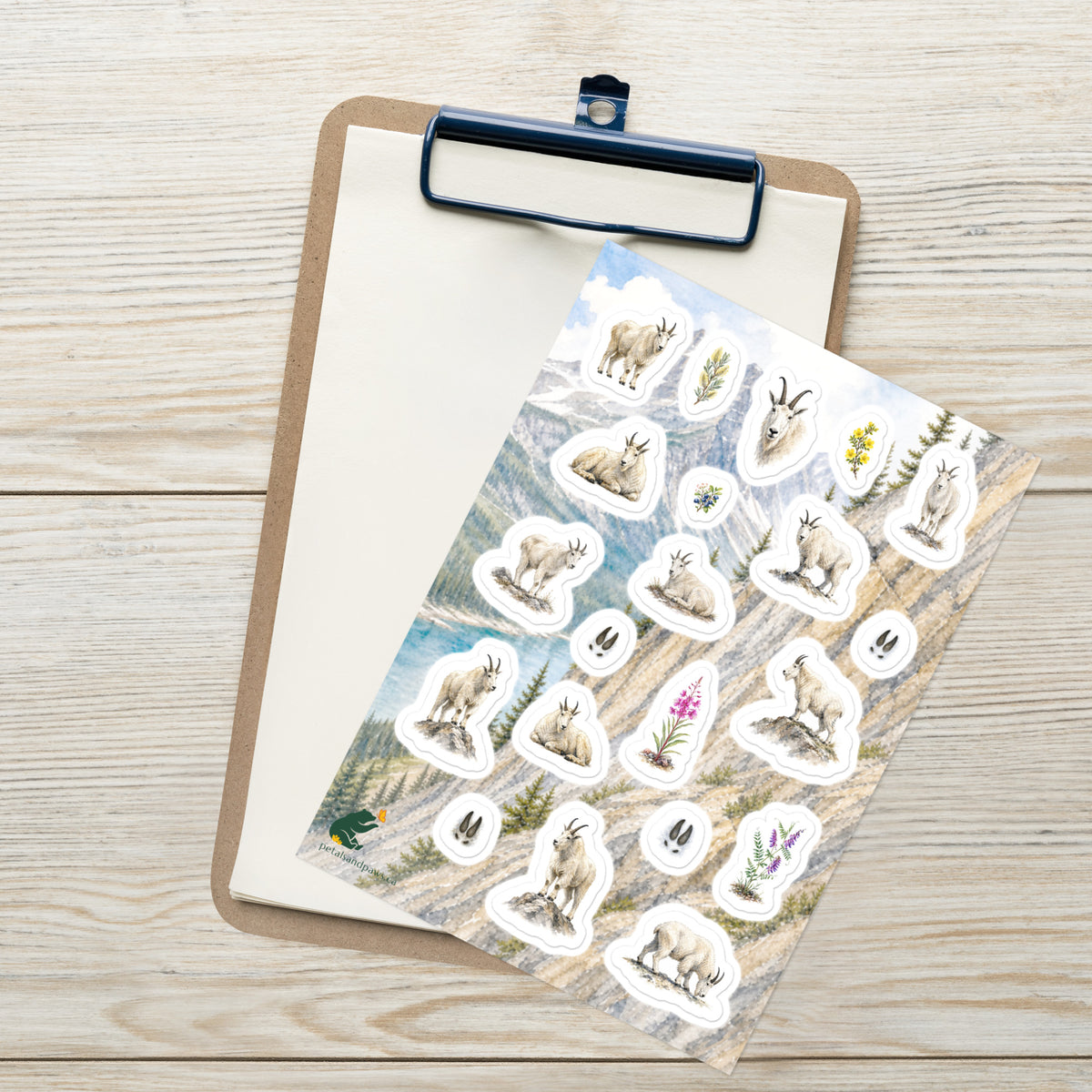 A sheet of kiss-cut stickers featuring illustrated mountain goat at rest, set against Rocky Mountain scenery, placed on top of an open clipboard with a blank sheet on a light wood tabletop.