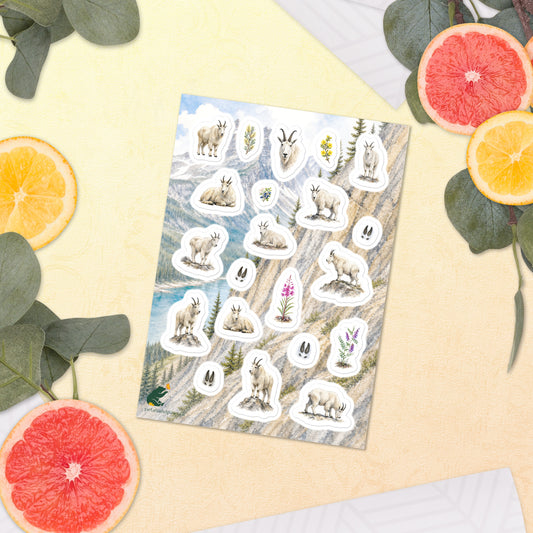 A sheet of kiss-cut stickers featuring Rocky Mountain landscape and mountain goat illustrations, placed next to citrus slices on a warm-colored background.