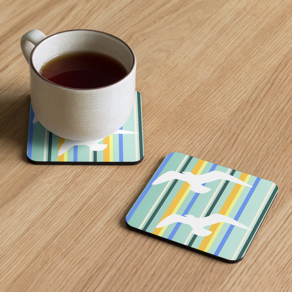 Product mockup of two Petals & Paws Cove Coasters, one with a mug of tea sitting on it, and the other set beside, both on a faux-wood surface. Design is seagulls are flying against a teal striped pattern.
