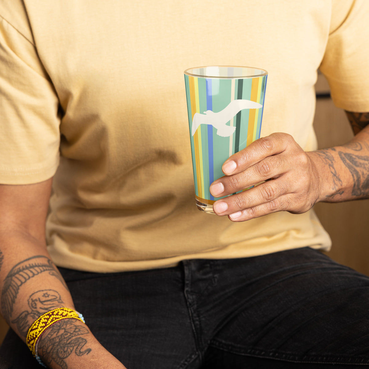 A man holding the Petals & Paws Cove Pint Glass, featuring seagull design with teal striped pattern. Chest and tattooed arms are within the picture.