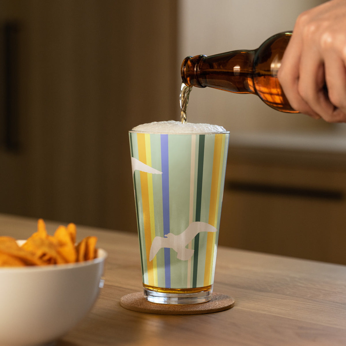 In-action mockup of Petals & Paws Cove Pint Glass, featuring seagull design with teal striped pattern. A hand is pouring beer into the pint glass and a bowl of nachos sits nearby.