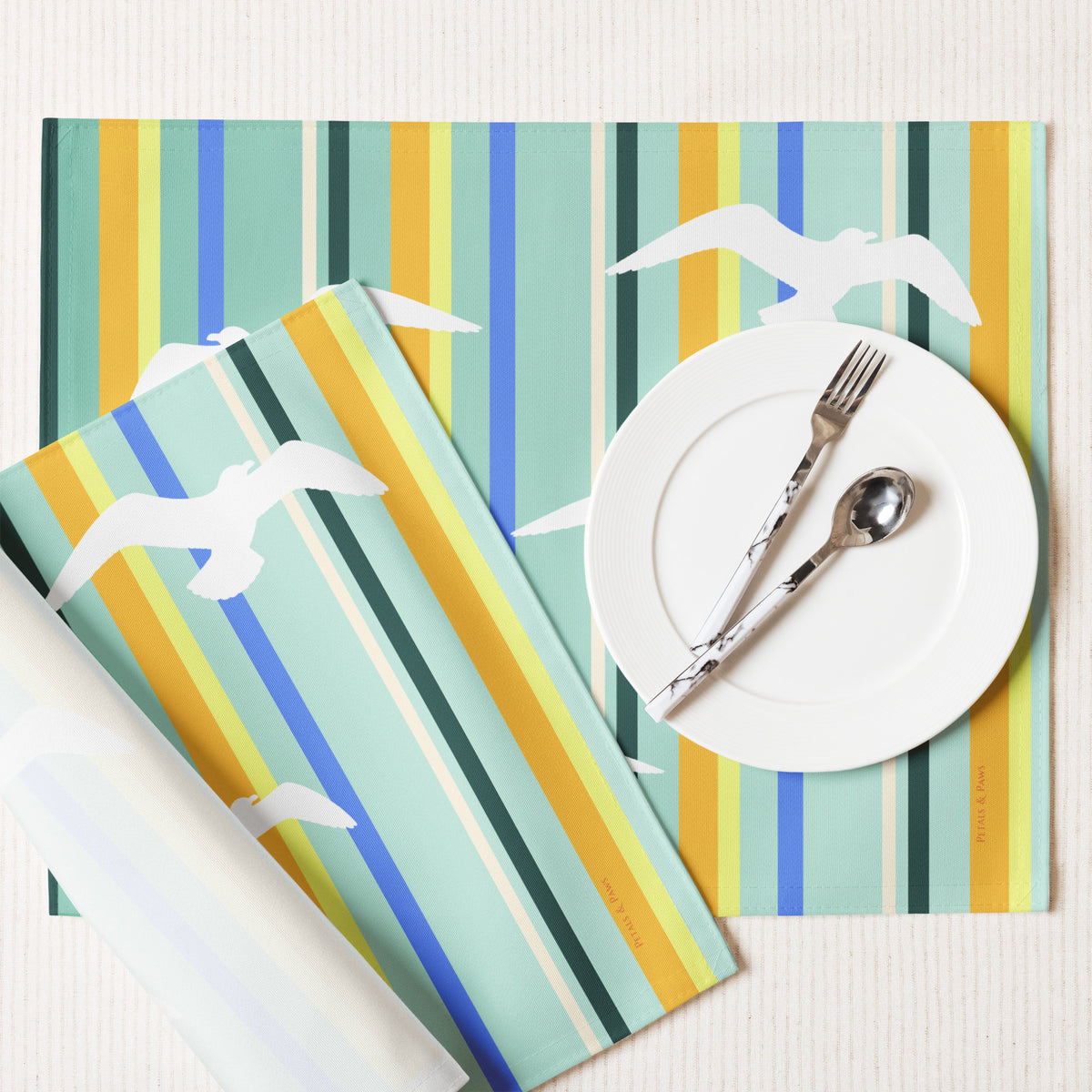 Product mockup of a Petals & Paws Cove Placemat, featuring seagull design with teal striped pattern. This image shows the white back and hemmed edges.