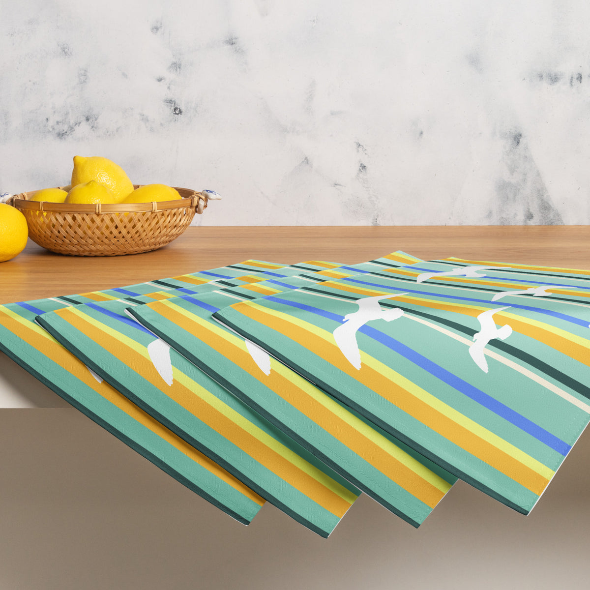 Product mockup of a Petals & Paws Cove Placemat, featuring seagull design with teal striped pattern. This image shows the full set of four with a basket of lemons.