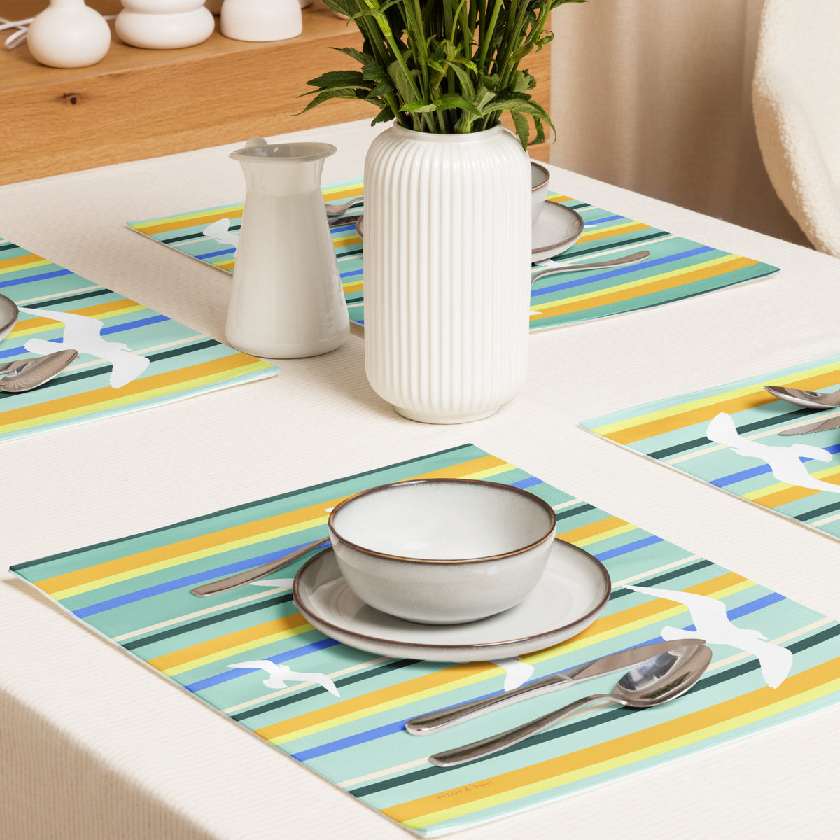 Product mockup of a Petals & Paws Cove Placemat, featuring seagull design with teal striped pattern. This image shows an overhead view of a dining table set for four with plates, bowls, utensils, and a vase holding cut flowers.