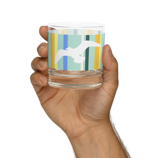 A Petals & Paws Cove Rocks Glass held up in a man's hand against a white background. This glass features a seagull design with teal striped pattern.