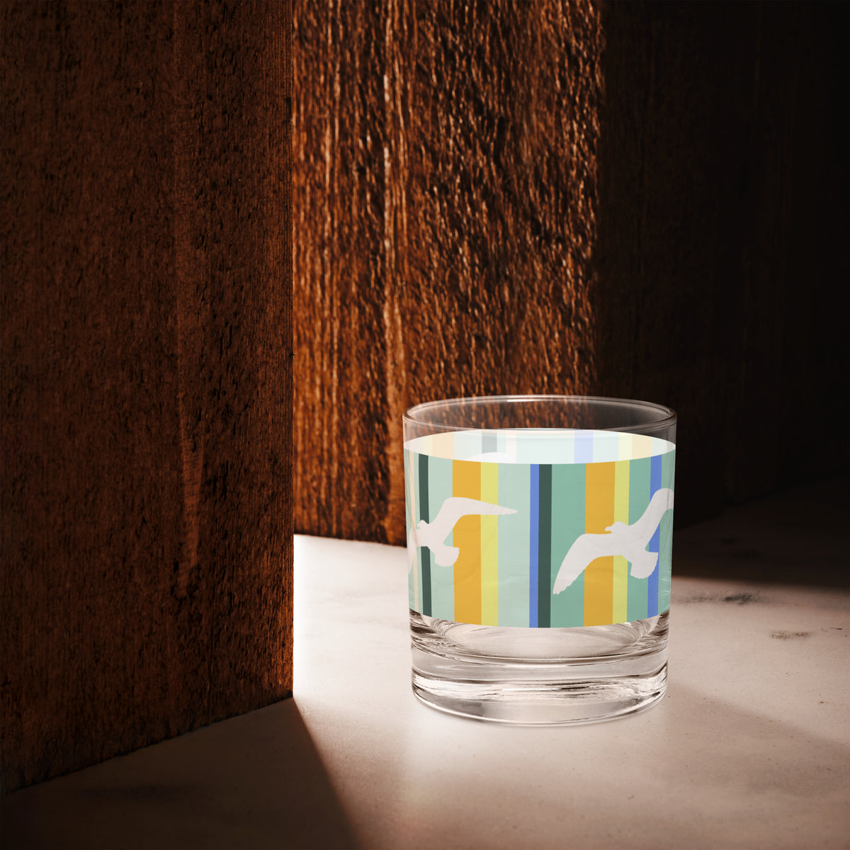 A Petals & Paws Cove Rocks Glass is sitting in the space of light between two cherry-wood-colored walls. This glass features a seagull design with teal striped pattern.