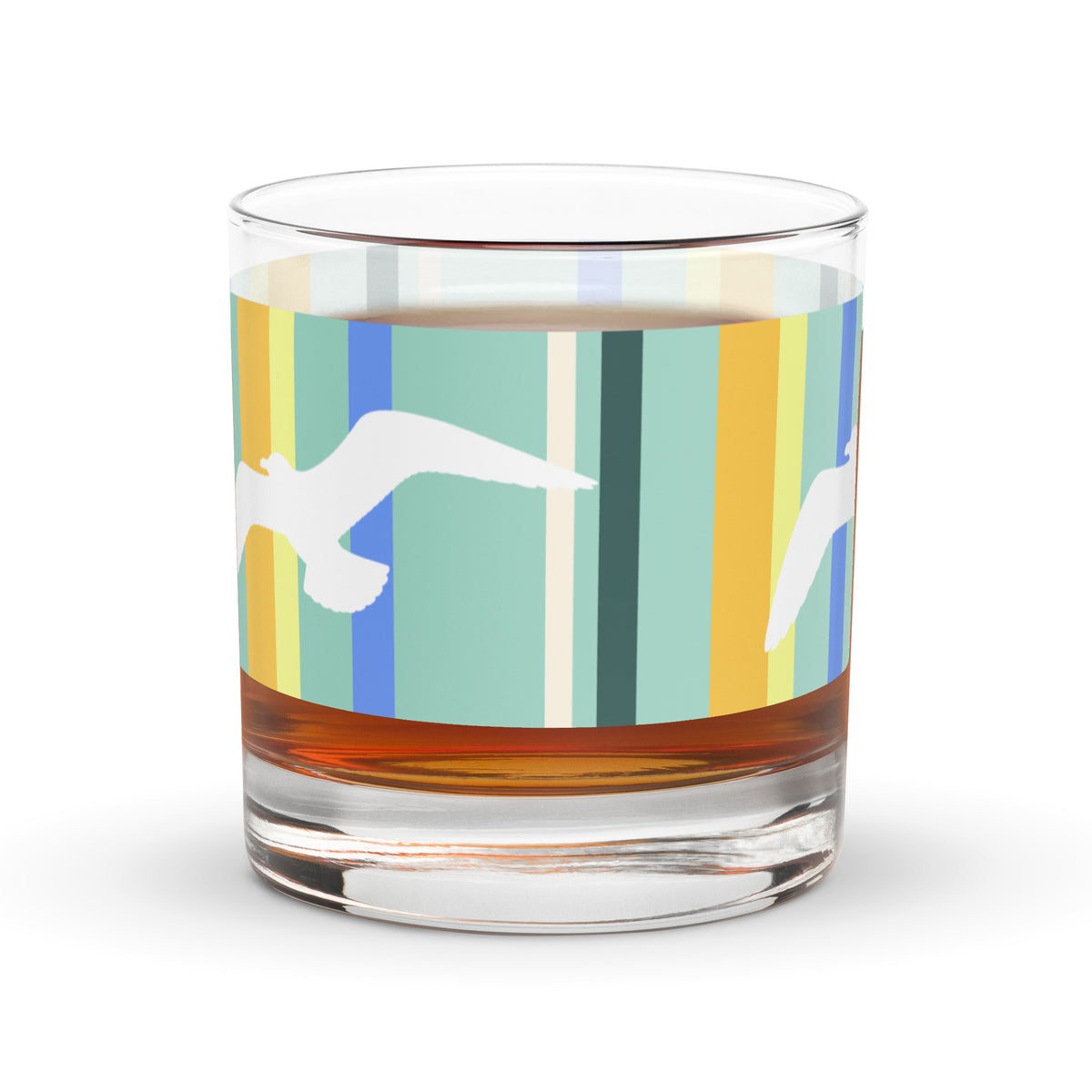 A Petals & Paws Cove Rocks Glass with a golden liquid inside it is sitting on a white surface. This glass features a seagull design with teal striped pattern.