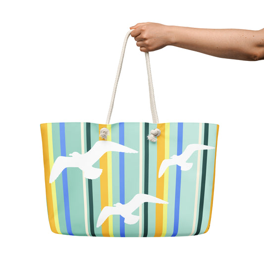 A Petals & Paws Cove Weekender Bag is held out against a white background. The female hand holds the bag by its cream-colored cotton rope handles. This all-over print bag features seagull design with teal striped pattern.