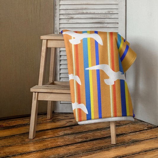 A 30 x 60" Petals & Paws Drift Beach Towel is draped on a wooden step stool against a neutral wall. This towel features seagull design with orange striped pattern.