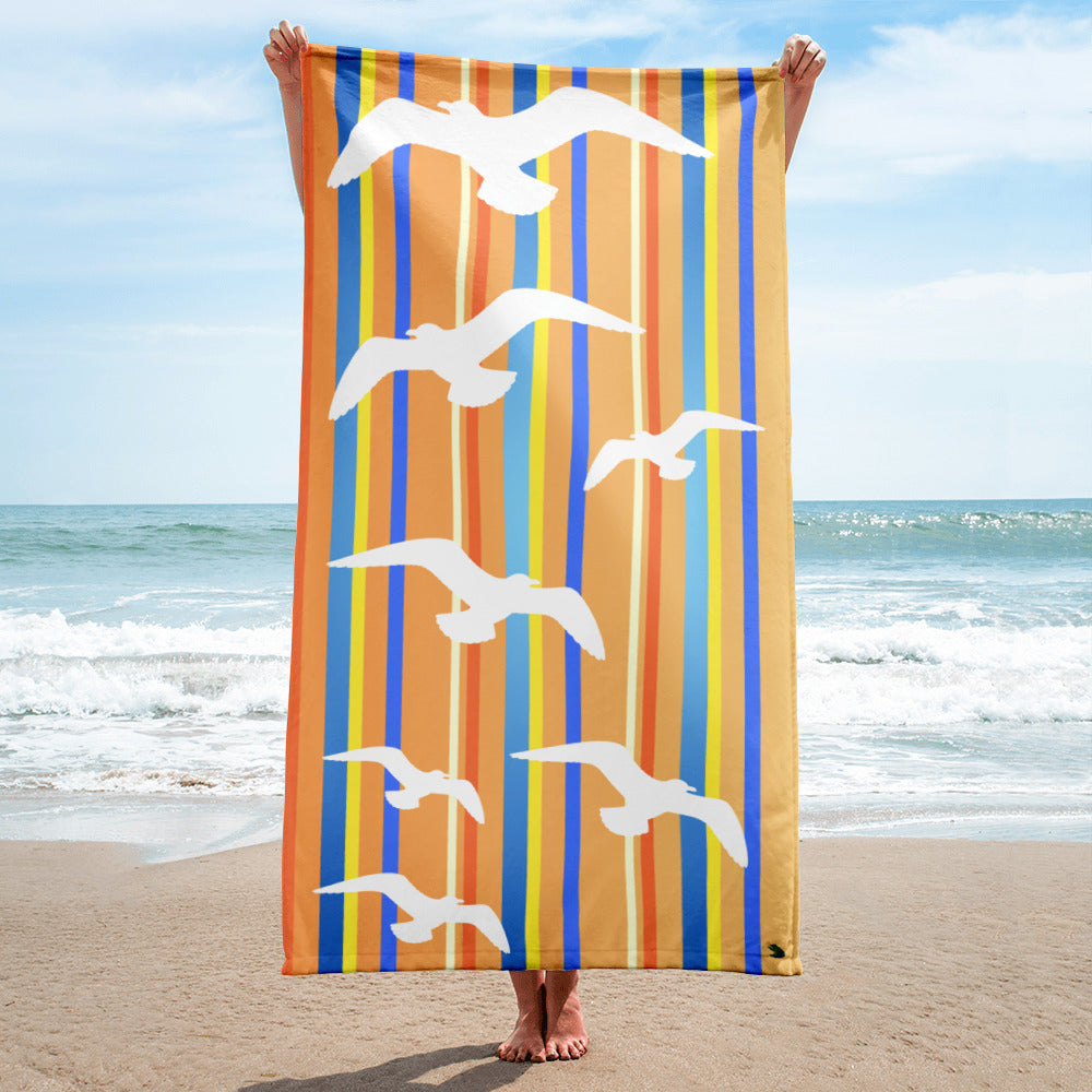 A 30 x 60" Petals & Paws Drift Beach Towel is held up vertically in front of a woman on a sandy beach. A large body of water is in the background. This towel features seagull design with orange striped pattern.